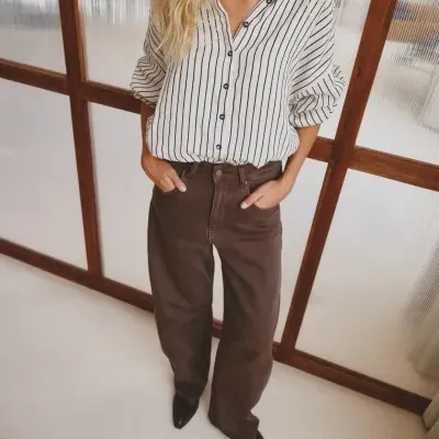 Mulher com camisa riscada branca e calças castanhas escuras num espaço interior