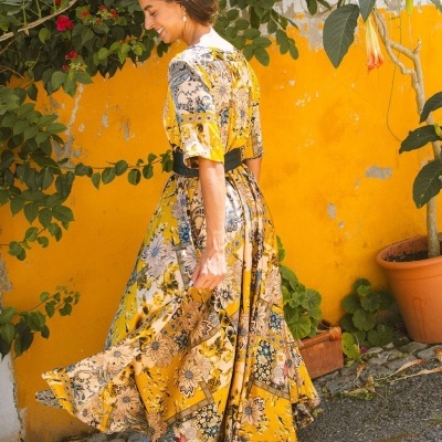 Vestido comprido floral amarelo com cinto preto e sandálias castanhas junto a parede amarela com plantas.