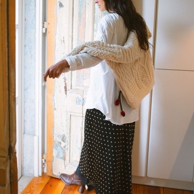 Mulher vestindo camisa branca, colete de malha creme, saia preta de bolinhas brancas e botas castanhas numa sala com chão de madeira.