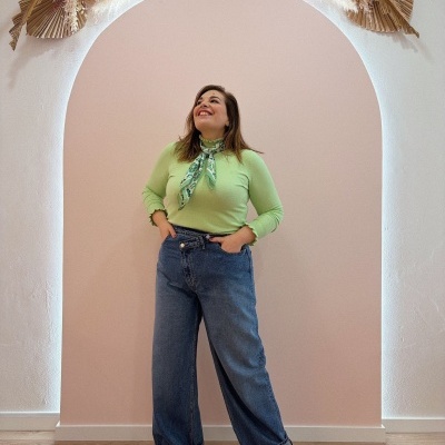 Mulher com camisola verde clara, lenço ao pescoço, jeans largos e ténis verdes em fundo rosa com arco decorado