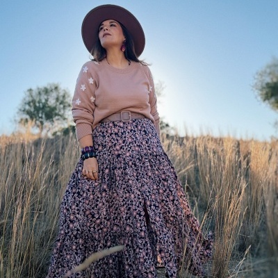 Mulher com suéter rosa, saia floral, chapéu marrom, pulseiras, em campo com céu azul