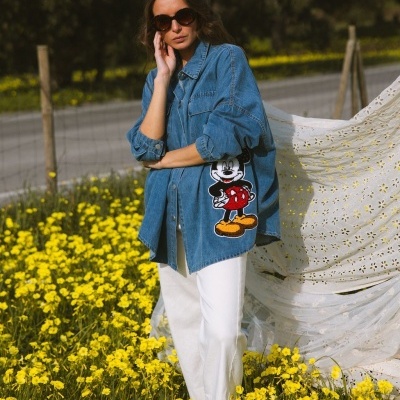 Camisa de ganga azul com estampado do Mickey Mouse, calças brancas e óculos escuros em campo de flores amarelas.