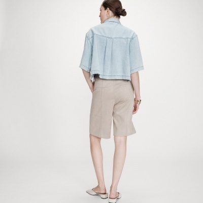 Modelo veste camisa jeans azul clara de manga curta e calções bege até ao joelho, sapatos mule brancos, fundo branco