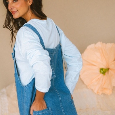 Mulher de costas com macacão de ganga azul claro e camisa branca em fundo neutro com almofada