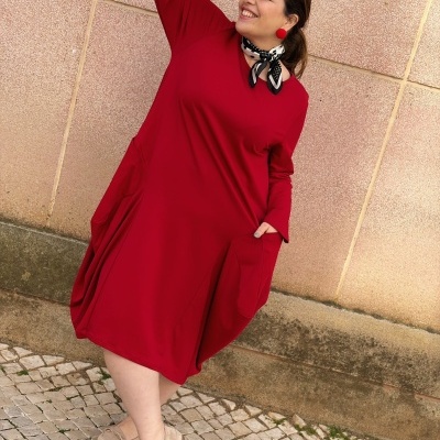 Mulher a vestir vestido vermelho com bolsos, lenço e brincos, sapatos beges e calçada de pedras