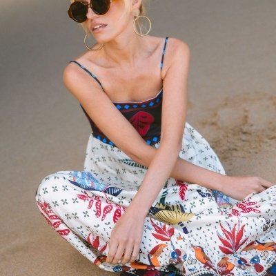 Mulher loira com vestido comprido colorido, óculos de sol redondos e brincos dourados sentada na areia