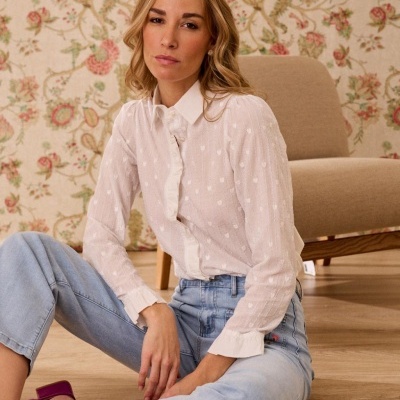 Blusa branca com folhos, jeans azul claro e sapatos rosa sentada em chão de madeira, cadeira bege e papel de parede floral ao fundo