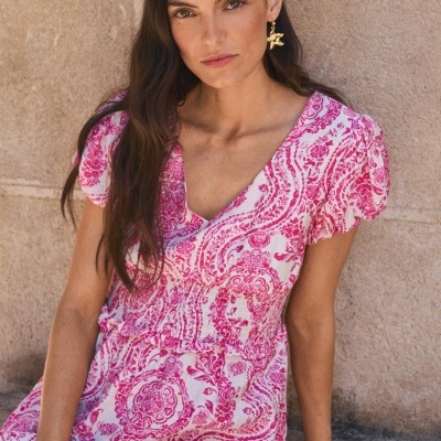 Mulher com vestido rosa e branco com padrão floral sentada junto a uma parede de pedra