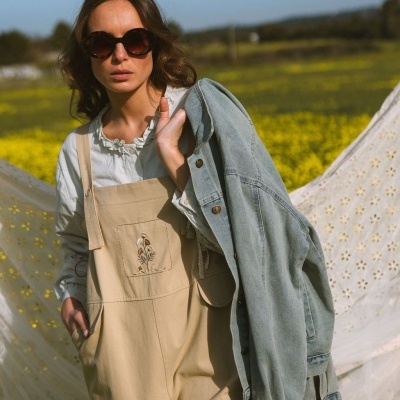 Mulher com óculos de sol, camisa branca, macacão bege com bordado e jaqueta de ganga azul claro