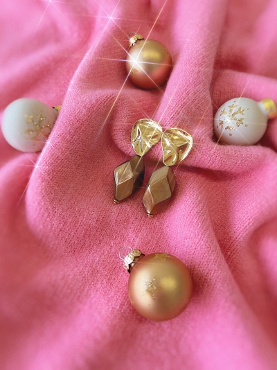 Brincos dourados geométricos sobre tecido rosa com bolas decorativas natalícias.