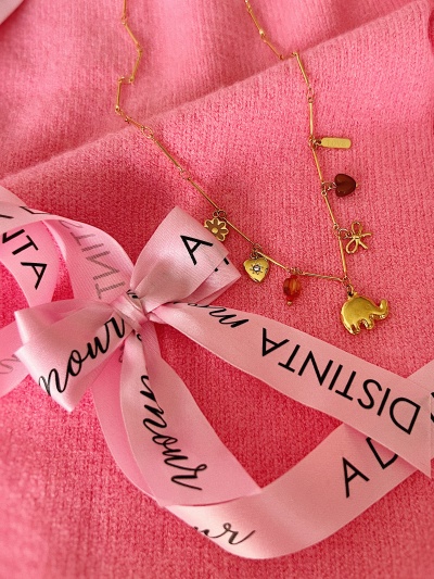 Colar dourado com pendentes sobre tecido rosa e fita rosa com texto.