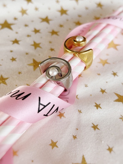 Dois anéis metálicos com pérolas em palhinhas cor-de-rosa e branca rodeados por fita rosa com texto