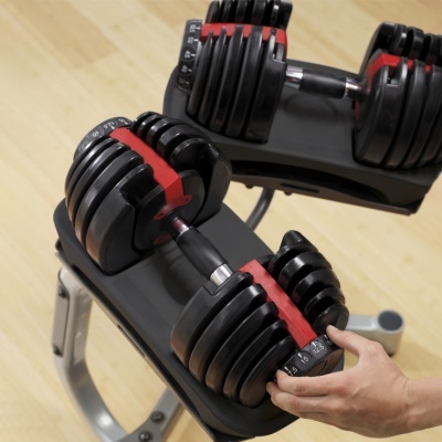 Halteres ajustáveis preto e vermelho em suporte cinza em ginásio, com chão de madeira clara.