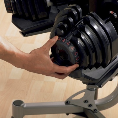 Halteres ajustáveis pretos com números vermelhos em suporte metálico com piso de madeira