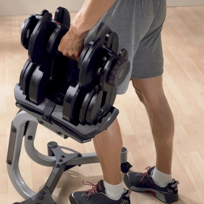 Halteres ajustáveis pretos numa base com pessoa de calções cinza de pé sobre piso de madeira.