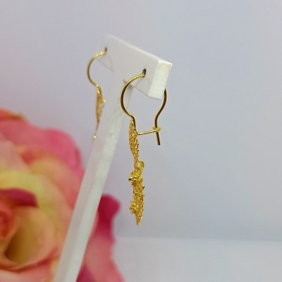 Brincos dourados pendentes exibidos em suporte branco com flor rosa ao fundo