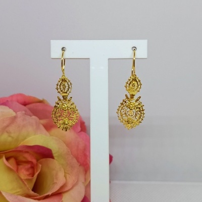 Brincos dourados pendentes com padrão rendado e flores, em suporte branco, flor colorida ao fundo