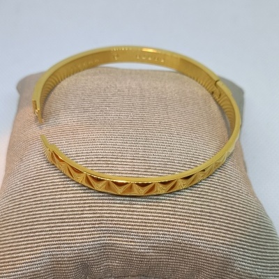 Pulseira tipo bracelete dourada com padrão geométrico sobre almofada bege