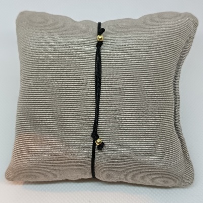 Pulseira com cordão preto e contas douradas sobre almofada cinza com linhas finas.