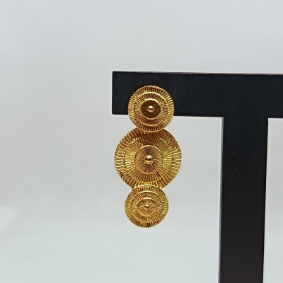 Brinco dourado com três discos texturados pendurado em suporte preto.