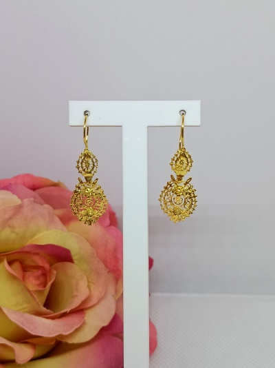 Brincos dourados pendentes com padrão rendado e flores, em suporte branco, flor colorida ao fundo