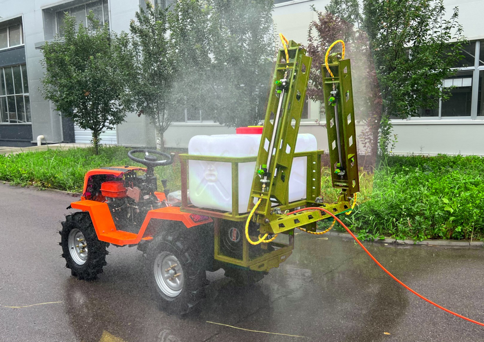 Trator agrícola com pulverizador e tanque a pulverizar líquido na rua junto de vegetação e edifícios.