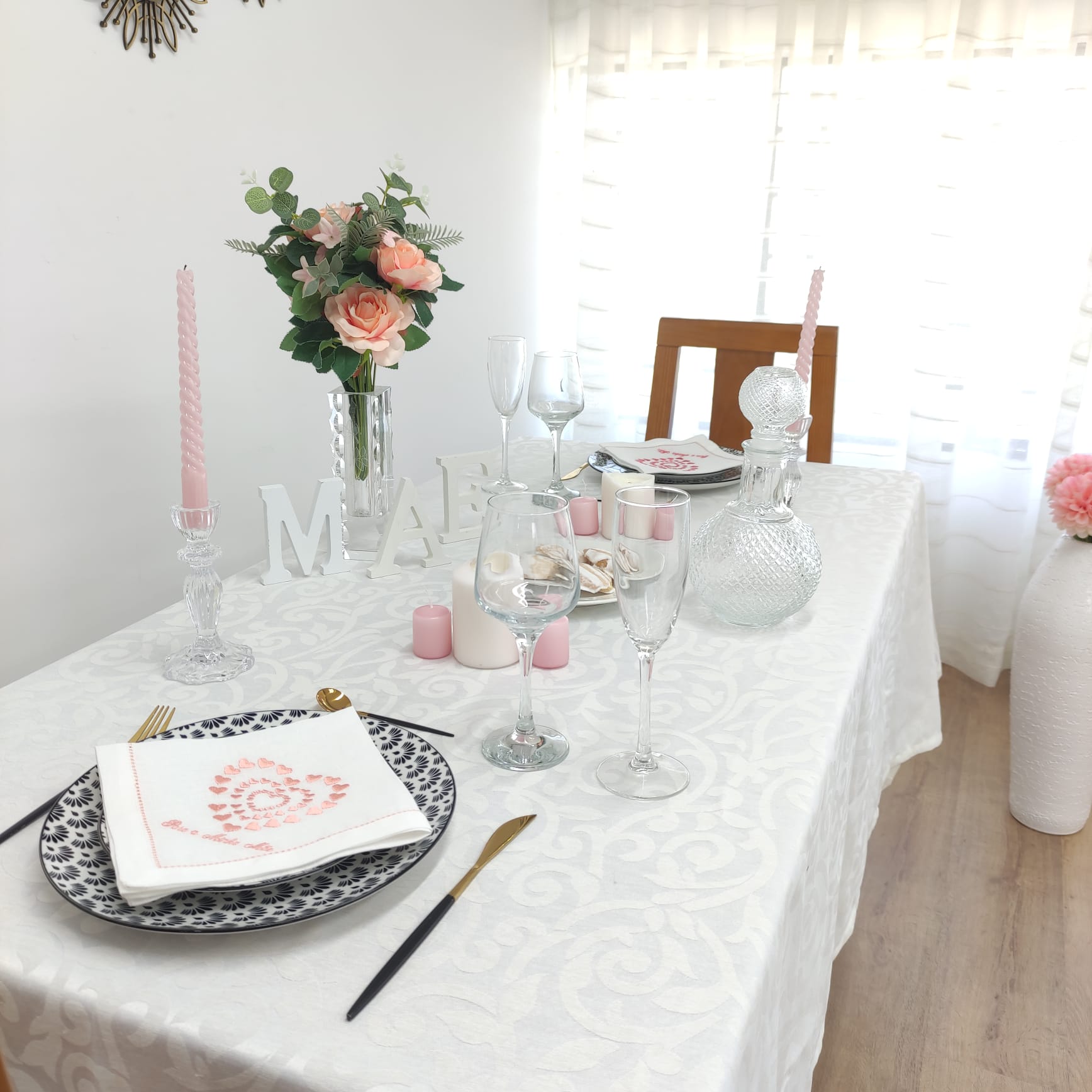 Mesa de jantar com toalha branca, pratos pretos com guardanapos rosa, talheres dourados e pretos, castiçais, jarra e vaso com flores.