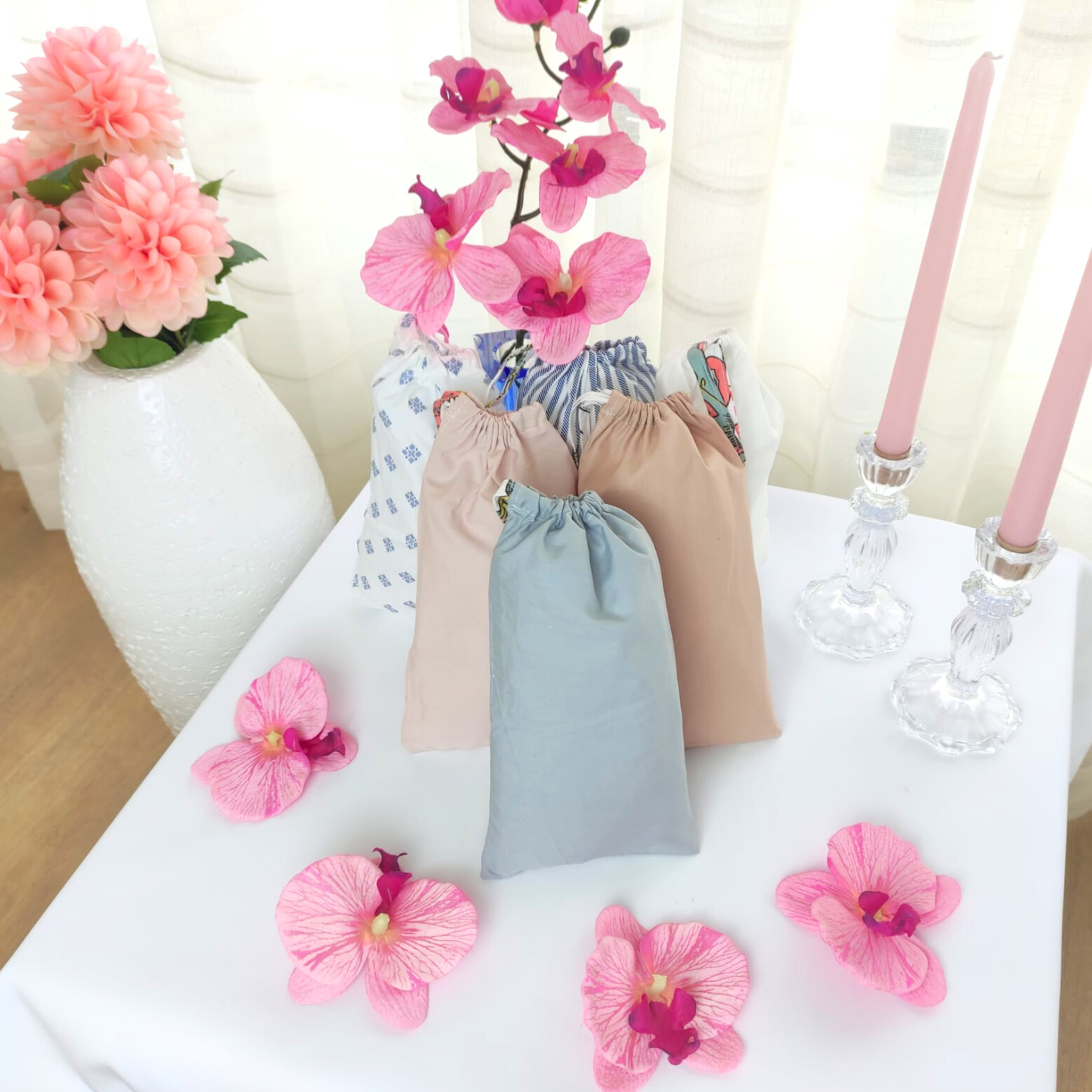 Quatro saquinhos de tecido em tons pastel sobre mesa branca com flores e castiçais de vidro