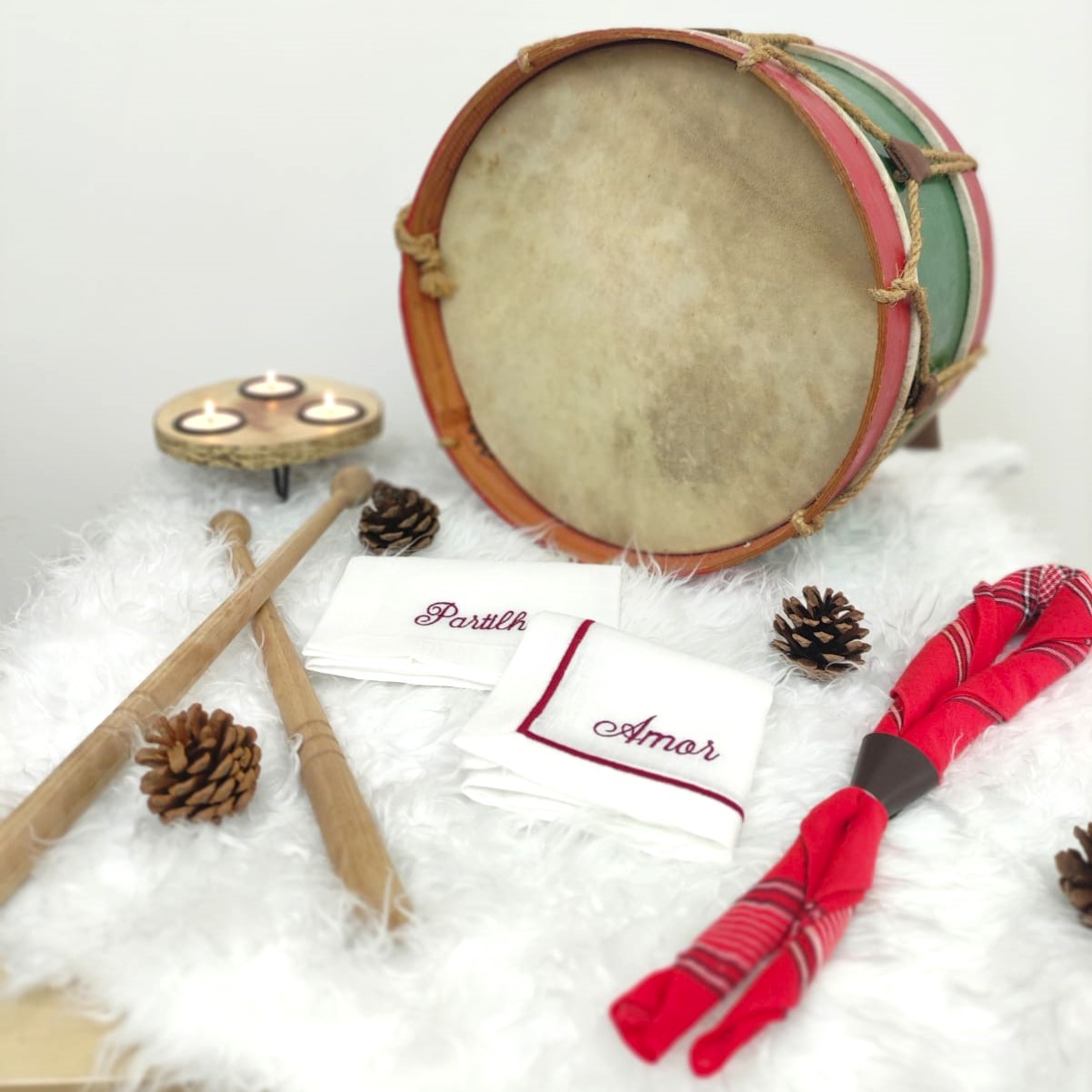 Tambor tradicional com palo de madeira, toalhas bordadas e velas sobre tecido felpudo branco