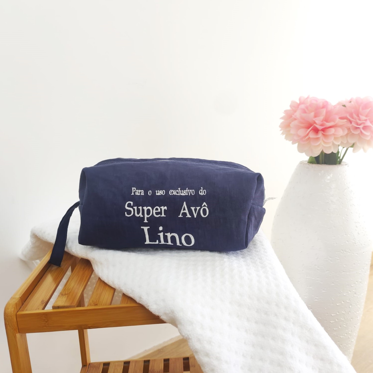 Necessaire azul com texto branco sobre toalha branca em estrutura de madeira, com vaso e flores ao lado.
