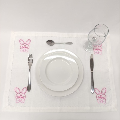 Mesa posta com louças brancas, talheres inox, copo de vidro e individual branco com bordados cor-de-rosa de coelhos