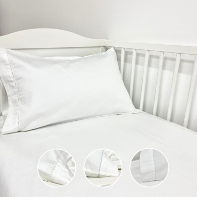 Conjunto de roupa de cama branco para berço com almofada e lençol, ambos em tecido liso, junto a berço branco.