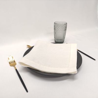 Mesa posta com prato preto, guardanapo branco, copo cinzento com textura, garfo dourado e faca preta.