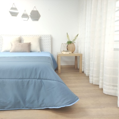 Quarto com cama azul, cabeceira branca, almofadas neutras, mesa de madeira com vaso e cortinas brancas