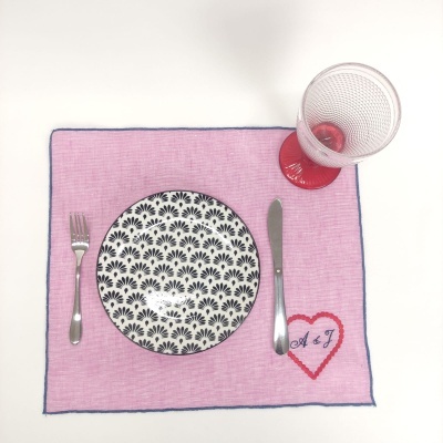 Jogo americano rosa com borda azul, prato decorado, talheres metálicos e copo com base vermelha