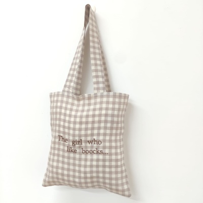 Saco de tecido padrão vichy bege e branco com texto bordado, pendurado numa parede