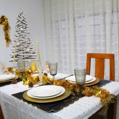 Mesa de jantar decorada com toalha branca, pratos brancos e dourados, copos cinzentos, decoração dourada e pinheiro branco no fundo