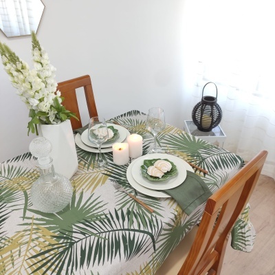 Mesa de jantar com toalha de folhas verdes e amarelas, pratos com guardanapos e flores, velas e jarra de vidro