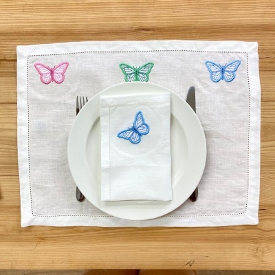 Jogo americano e guardanapo branco bordados com borboletas coloridas sobre mesa de madeira clara