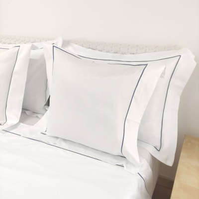 Jogo de almofadas e lençóis brancos com acabamento azul numa cama com cabeceira branca e mesa de cabeceira clara