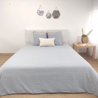 Cama arrumada com roupa de cama azul riscada e almofadas, mesa de cabeceira em madeira com vaso, espelhos hexagonais na parede.
