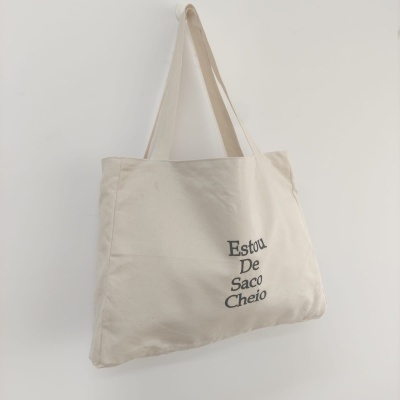 Saco de tecido bege claro com alças e texto bordado preto pendurado numa parede branca