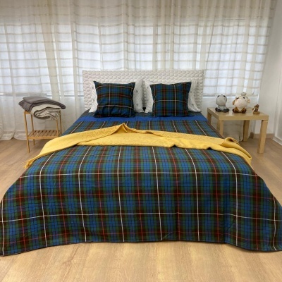 Conjunto de cama com colcha xadrez em vários tons, almofadas combinando, cobertor amarelo, criado-mudo de madeira com objetos e cortinas brancas.