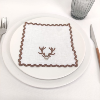 Guardanapo branco com borda ondulada castanha e bordado de rena em prato branco com talheres e copo cinzento