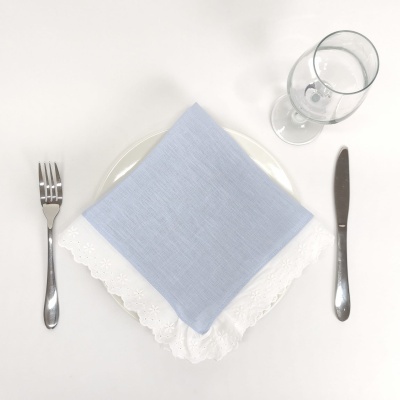 Conjunto de mesa com talheres de aço inoxidável, prato branco com guardanapos azul claro e branco bordado, e taça de vidro sobre mesa branca.