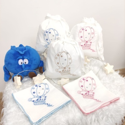 Saquinhos brancos bordados com balão e panos de boca com bordado correspondente, junto a brinquedo azul em superfície fofa branca.