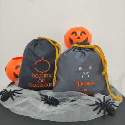 Duas saquetas cinzentas com cordão amarelo e escrita laranja em cima de rede branca com aranhas e abóboras de Halloween