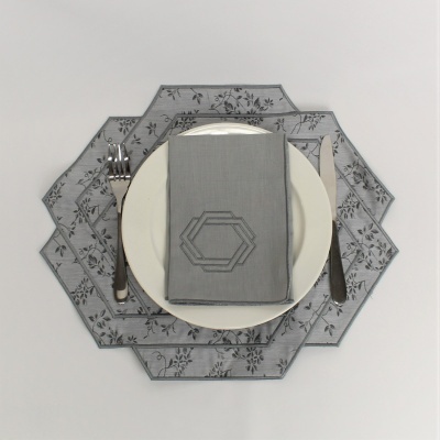 Conjunto de loiça com prato branco, base de tecido cinza com padrão floral, guardanapo cinza com bordado e talheres prateados