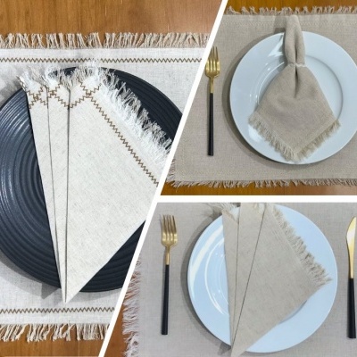 Guardanapos beje com franjas e costura castanha sobre pratos com talheres dourados e cabos pretos numa mesa de madeira.