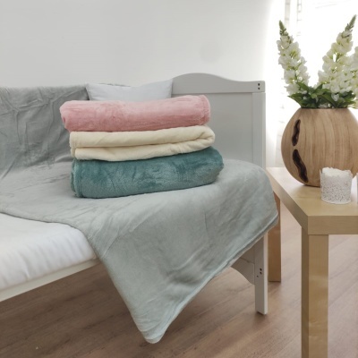 Três mantas felpudas empilhadas em tons rosa, creme e azul-petróleo sobre cama com manta cinza, ao lado mesa com vaso e flores brancas